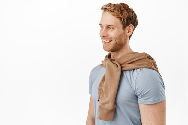 Happy candid man with strong body and red hair, looking left and standing half turned as if talking to someone, smiling pleased, having conversation, standing in casual clothes over white background