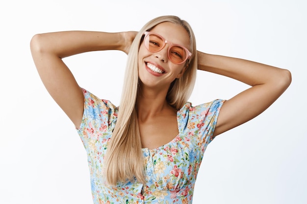 Happy and carefree blond girl in sunglasses holding hands behind head resting and laying back standing over white background