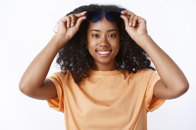 Happy carefree stylish modern millennial dark-skinned woman take off glasses smiling broadly feeling enjoyment relaxing sunbathing travelling hot country, standing pleased white wall