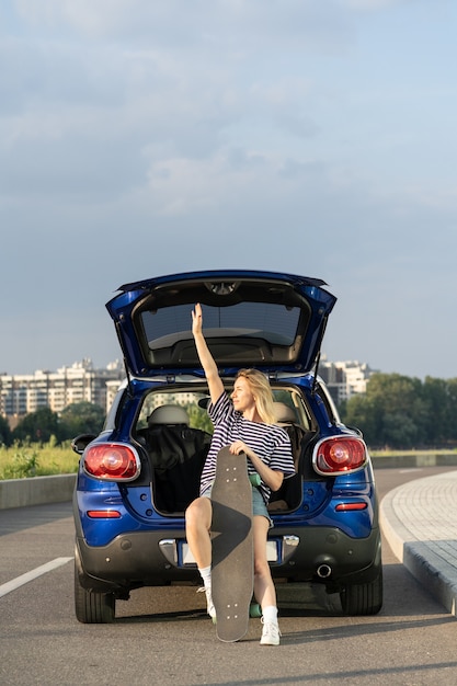 Happy carefree woman travel by car sit in trunk with longboard enjoy sunset during summer vacation