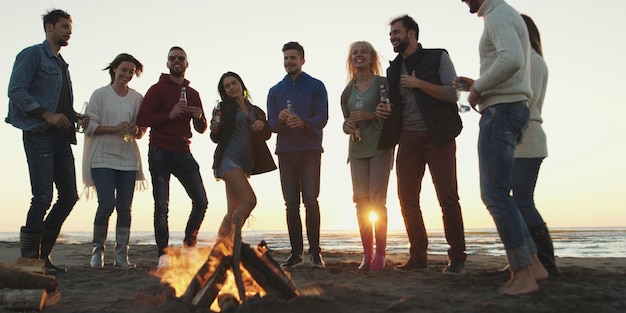 Happy Carefree Young Friends Having Fun And Drinking Beer By Bonefire On The Beach As The Sun Begins To Set