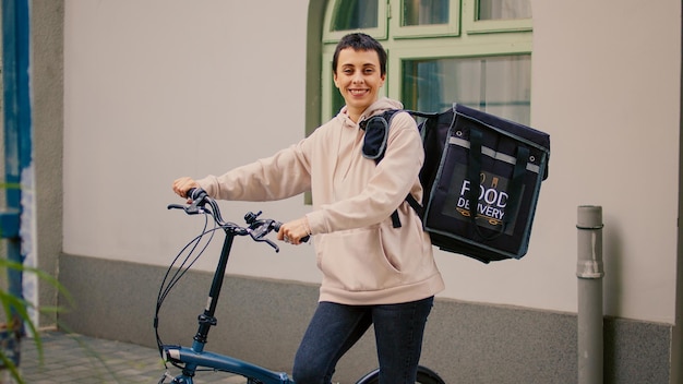 Happy carrier sitting at front door to deliver takeaway meal\
order, carrying backpack with lunch food. delivery service female\
worker delivering fastfood in bag and riding bicycle.