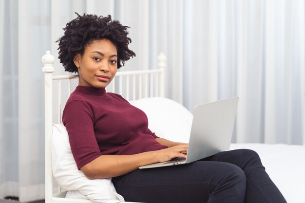 Happy casual beautiful american african woman working on laptop computer while setting  on the bed in the house.