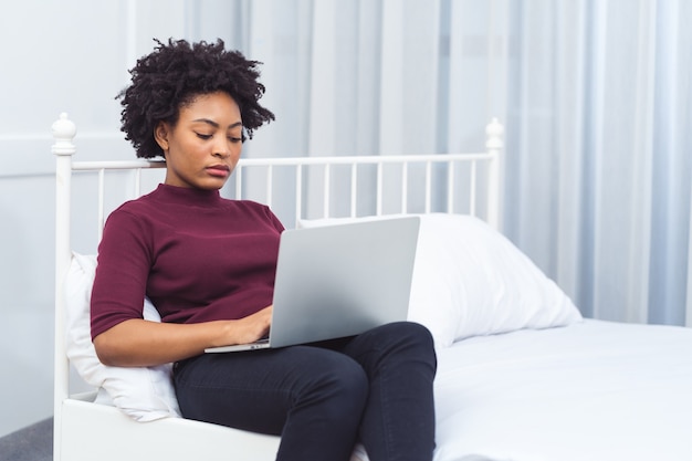 Happy casual beautiful american african woman working on laptop computer while setting  on the bed in the house.