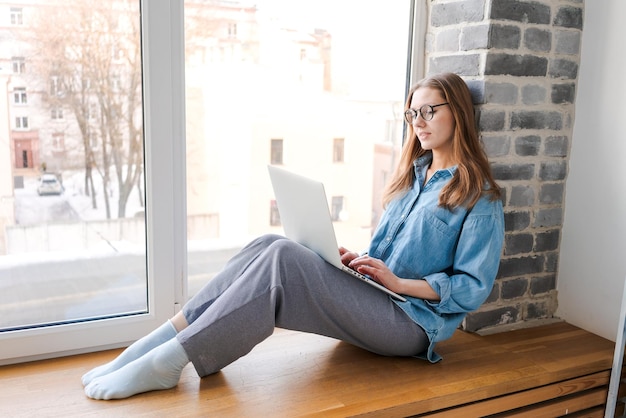 Счастливая случайная красивая женщина в очках, работающая на ноутбуке, сидящая на подоконнике