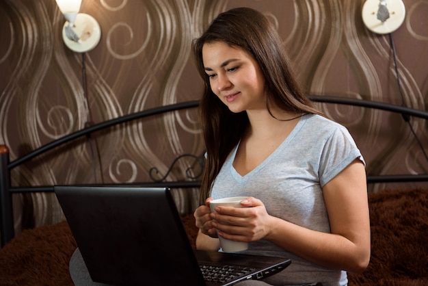 Bella donna casuale felice che lavora ad un computer portatile che si siede sul letto in casa.