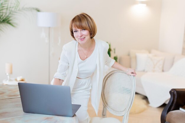 Happy casual beautiful woman working on a laptop sitting on the chair in the stylish white living room