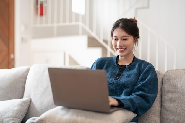 家のソファに座ってラップトップに取り組んで幸せなカジュアルな美しい若いアジアの女性