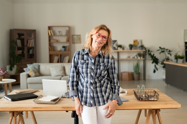 Happy casual blond female of middle age in her workplace with table, laptop, supplies and documents at home