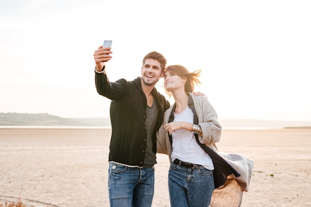 Happy casual couple making selfie photo while standing together on the beach at sunlight