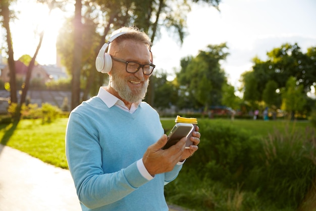 Happy casual senior man wearing headset having video conference or watching funny movie on mobile drinking coffee on walk in public park