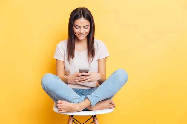 Happy casual woman sitting on the chair and using smartphone over yellow wall