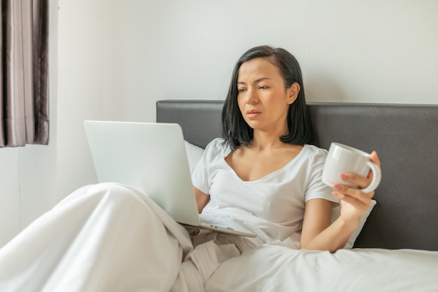 Happy casual woman working on a laptop sitting on the bed in the house.