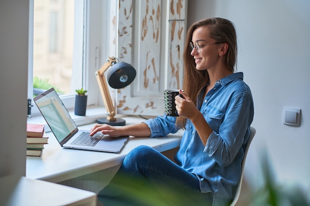 Счастливая случайная молодая улыбающаяся женщина в круглых очках с чашкой кофе, просматривающая онлайн-страницу за компьютером на уютном, удобном домашнем рабочем месте у окна