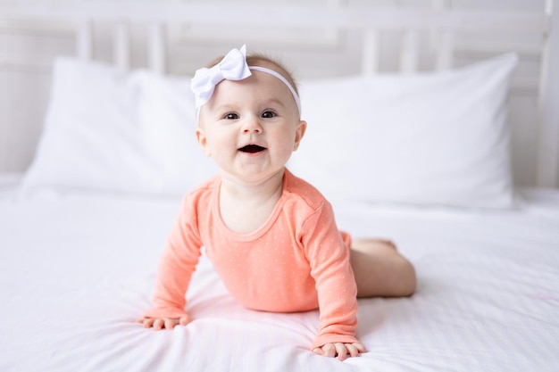 A happy Caucasian baby girl in a pink bodysuit lies on the bed at home in the bedroom on white bedding looks at the camera laughs a healthy baby
