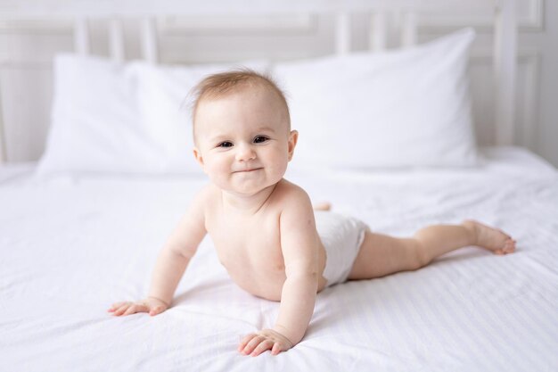 Happy caucasian baby girl in white panties lies on her stomach\
on the bed at home in the bedroom on white bed linen looks at the\
camera laughs healthy baby