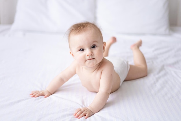 Happy caucasian baby girl in white panties lies on her stomach\
on the bed at home in the bedroom on white bed linen looks at the\
camera laughs healthy baby