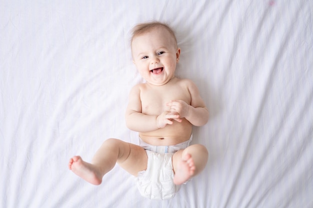 Happy Caucasian baby girl in white panties lies on her stomach on the bed at home in the bedroom on white bed linen looks at the camera laughs healthy baby