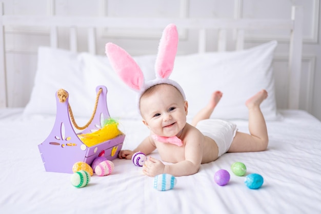 A happy Caucasian baby girl with a hare ears rim on her head and in panties lies on the bed at home with colorful Easter eggs and yellow spring flowers looks at the camera laughs Easter baby