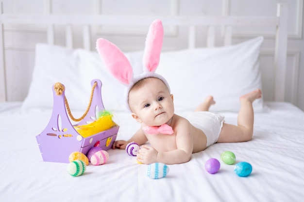 A happy Caucasian baby girl with a hare ears rim on her head and in panties lies on the bed at home with colorful Easter eggs and yellow spring flowers looks at the camera laughs Easter baby
