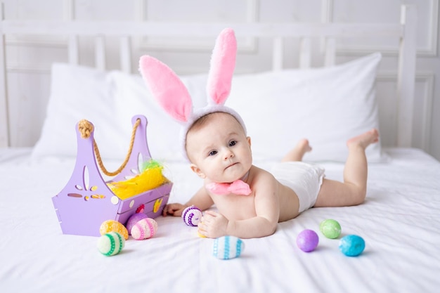 A happy caucasian baby girl with a hare ears rim on her head and in panties lies on the bed at home with colorful easter eggs and yellow spring flowers looks at the camera laughs easter baby