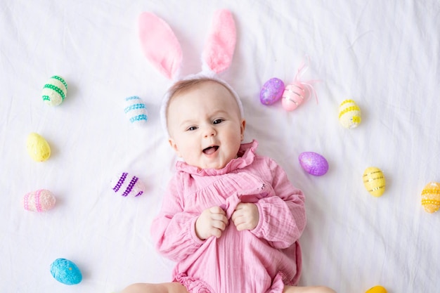 A happy Caucasian baby girl with a rim of bunny ears on her head lies on her back on the bed at home in the bedroom with colorful Easter eggs looks at the camera laughs Easter baby