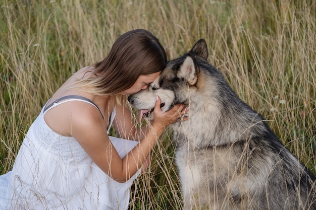 Happy Caucasian blonde woman in white dress hugging and kiss alaskan malamute dog in summer field. face to face. love and friendship between human and animal.