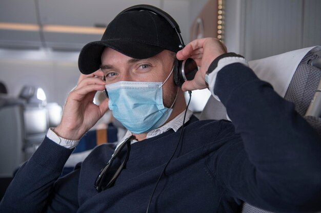 写真 ハッピーな40代の白人ビジネスクラスの乗客 マスクをかぶってヘッドフォンで 飛行機内で音楽を聴いて映画を見る