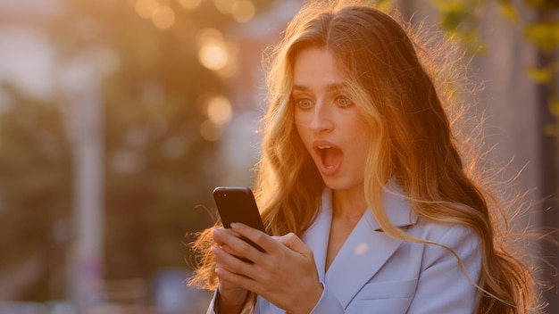 Happy caucasian business woman enjoy success looking in mobile\
phone in city sun background closeup joyful girl reading good news\
in smartphone makes yes hand gesture lady celebrating online\
victory