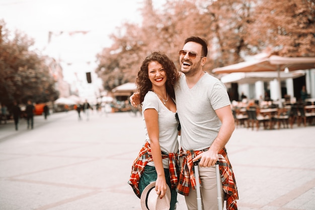 Happy Caucasian couple standing on the street and posing with hat and luggage in hands. Traveling concept.