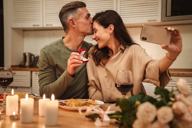 happy caucasian couple taking selfie photo with engagement ring while having romantic candlelight dinner at home