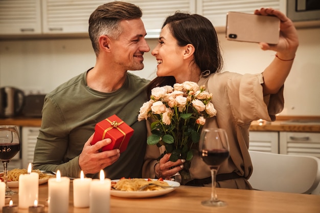 happy caucasian couple taking selfie photo with flowers while having romantic candlelight dinner at home