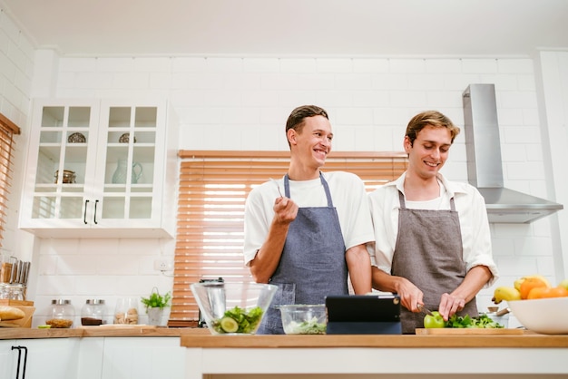 幸せな白人のゲイカップルがタブレットで料理の方法を見ながらキッチンで一緒に料理しています