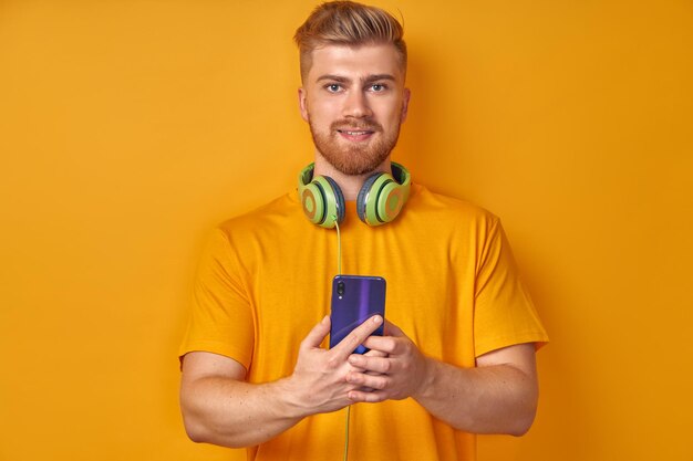 Happy caucasian ginger man enjoys music in headphones holds modern cell phone on orange background