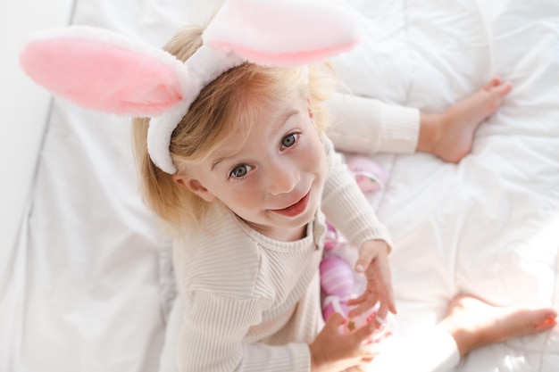 Happy caucasian girl two years old wearing rabbit ears playing with eggs sitting on bed looking at c