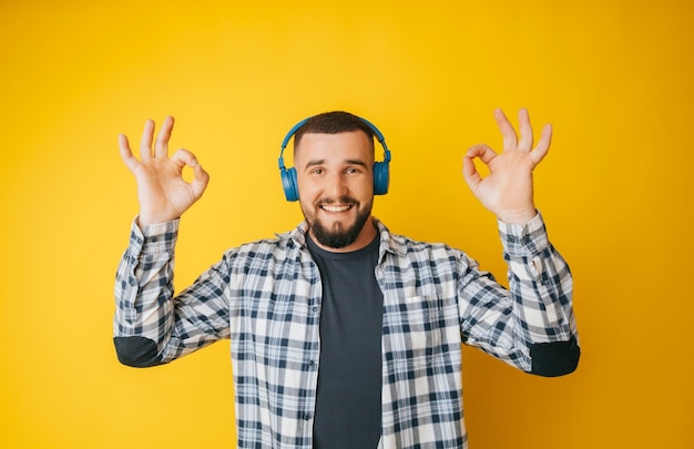 Happy caucasian guy shows a good gesture agrees with something claims that everything is fine smiles broadly wears headphones enjoys isolated on a yellow background