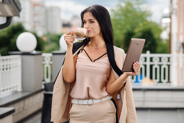 Happy caucasian lady in suit with tablet and holding a cup of coffee outdoors the hotel terrace