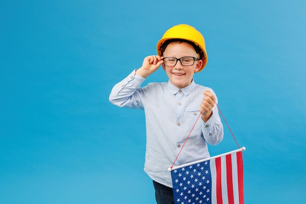 Happy Caucasian little child boy in yellow safety helmet holding and waving American flag with smiling face