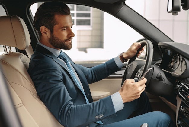 Happy caucasian male in formal wear driving an automobile and holding smartphone in hand