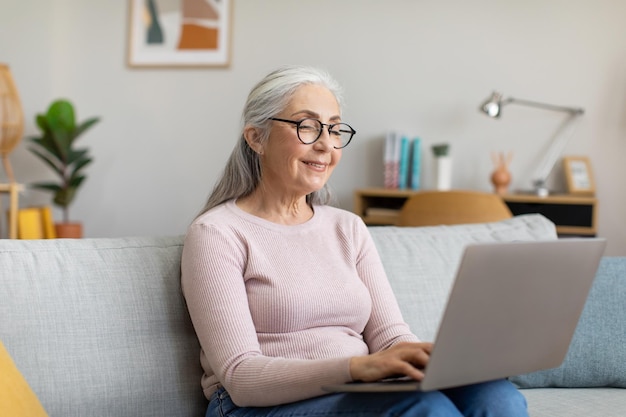 Happy caucasian mature grayhaired female in glasses typing on laptop watching video in living room interior