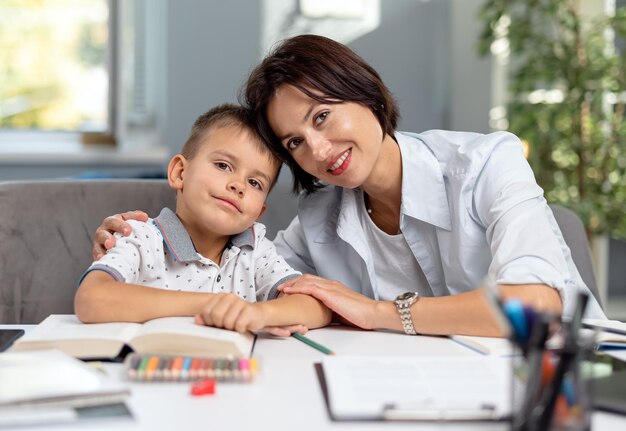 Happy caucasian mother embracing her lovely son while sitting together at desk with books and notes Domestic studying and family time concept