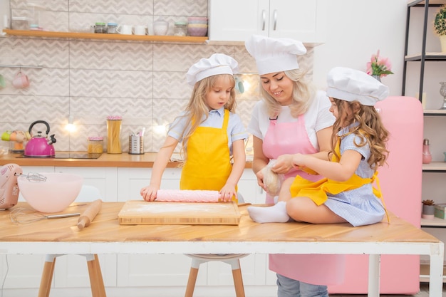 Happy caucasian mother helping her preschool daughters rolling dough for homemade bakery. Two female generations family in cooking process together in kitchen.