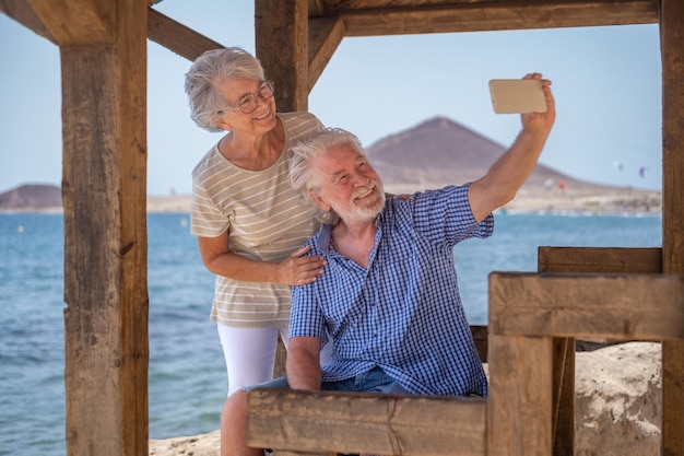 Happy caucasian senior couple sitting in the shade of the\
gazebo in front of the sea looking at mobile phone for a selfie\
elderly pensioners enjoying sea vacation in a windy sunny day