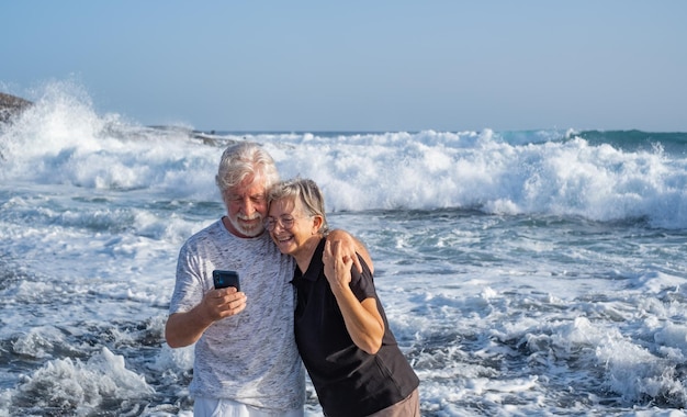 夕日の海辺に立つ幸せな白人老夫婦が携帯電話を見て微笑み、自由時間の海での休暇や退職を楽しんでいる年金受給者