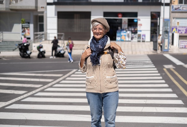 밝은 도시 중심가에서 길을 건너는 행복한 백인 노인 여성은 배낭을 들고 긍정적인 기분을 표현합니다.