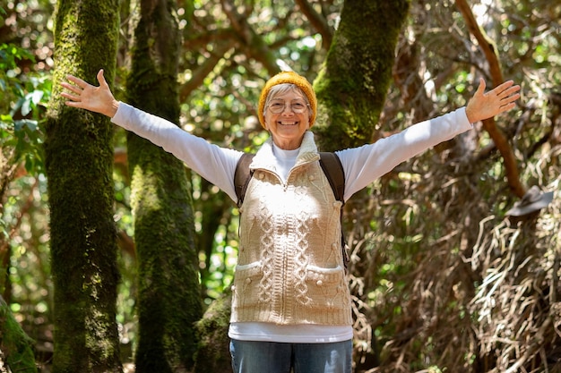 Photo happy caucasian senior woman hiking in the forest holding backpack enjoying freedom and nature elderly lady with eyeglasses and outstretched arms