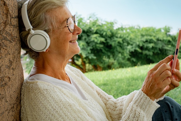 Happy caucasian senior woman sitting in public park lawn leaning against a tree trunk while using mobile phone