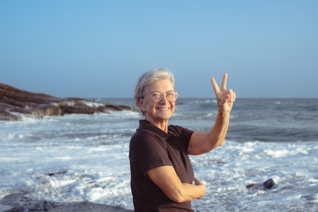 해질녘 해변에 서 있는 행복한 백인 노인 여성은 바다와 푸른 하늘 위로 지평선 위로 엄지손가락으로 카메라를 바라보고 있다