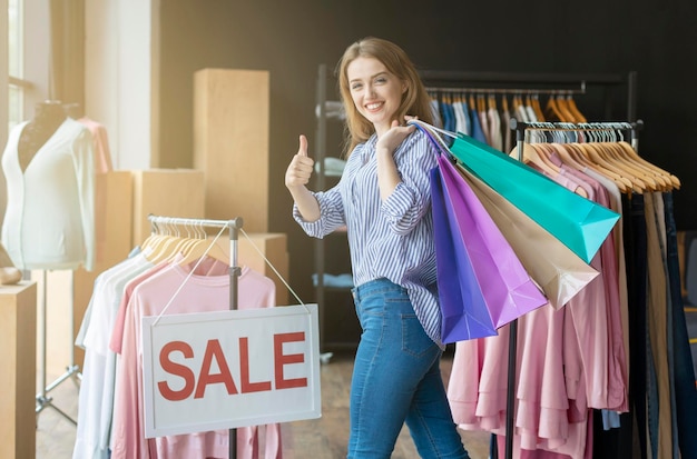 Happy caucasian shopper holding shopping bags on shoulder at boutique, showing thumb up, sale board