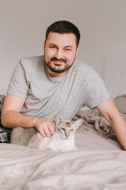 Foto felice uomo barbuto caucasico sorridente seduto sul letto nella camera da letto a casa e accarezzando accarezzare il gatto